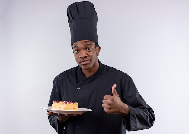 Heureux jeune cuisinier afro-américain en uniforme de chef tient le gâteau sur la plaque et les pouces vers le haut isolé sur mur blanc