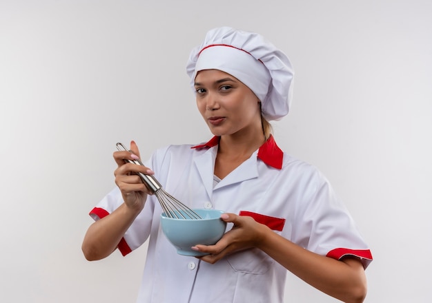 Heureux jeune cuisinier femme portant l'uniforme de chef tenant un fouet et un bol sur un mur blanc isolé