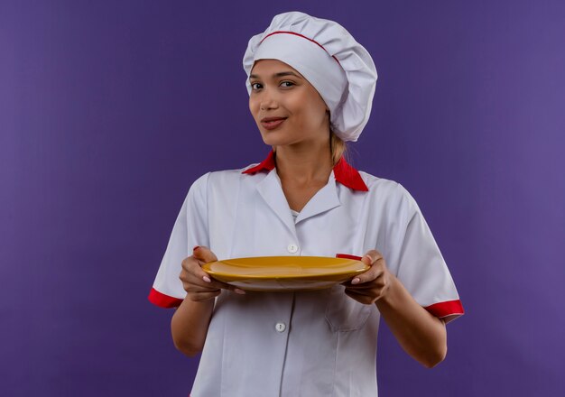 Heureux jeune cuisinier femme portant l'uniforme de chef tenant la plaque sur un mur isolé avec copie espace