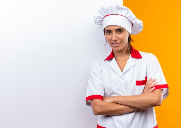 Heureux jeune cuisinier portant l'uniforme de chef debout avec un mur blanc et croisant les mains avec copie espace