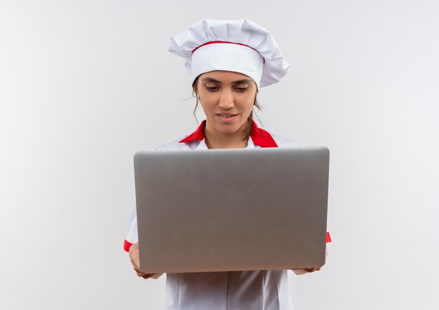 Heureux jeune cuisinier portant l'uniforme de chef tenant et à la recherche d'un ordinateur portable sur un mur blanc isolé avec copie espace