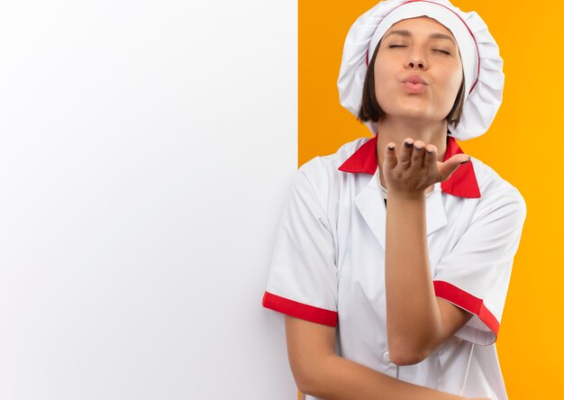 Heureux jeune cuisinier en uniforme de chef debout devant un mur blanc et envoi d'un coup de baiser à l'avant avec les yeux fermés isolé sur un mur orange