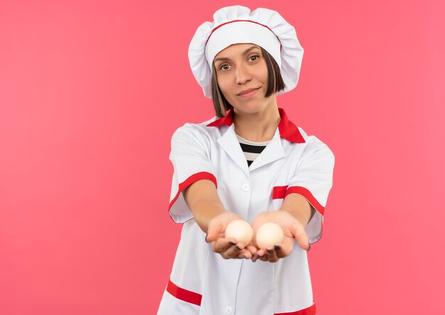 Heureux jeune cuisinier en uniforme de chef étendant les œufs vers l'avant isolé sur mur rose