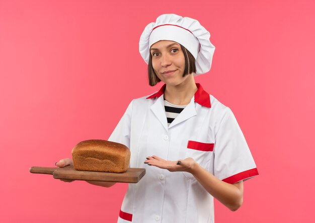 Heureux jeune cuisinier en uniforme de chef tenant et pointant avec la main sur une planche à découper avec du pain dessus isolé sur fond rose