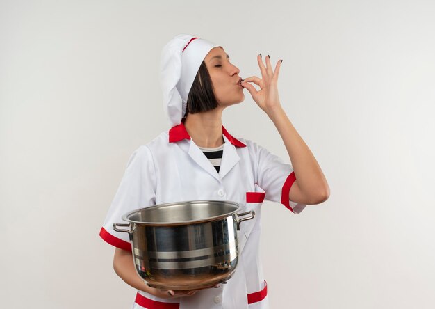 Heureux jeune cuisinier en uniforme de chef tenant le pot et faisant un geste savoureux avec les yeux fermés isolé sur un mur blanc