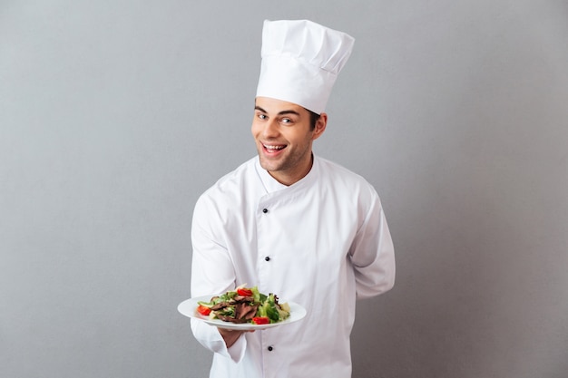 Photo gratuite heureux jeune cuisinier en uniforme tenant la salade.