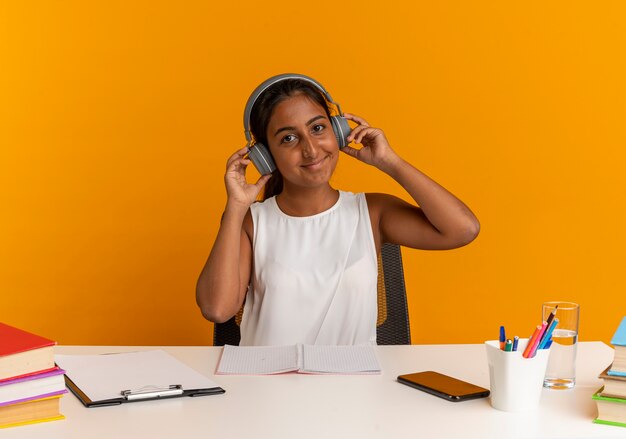 Heureux jeune écolière assis au bureau avec des outils scolaires écouter de la musique sur un casque isolé sur un mur orange