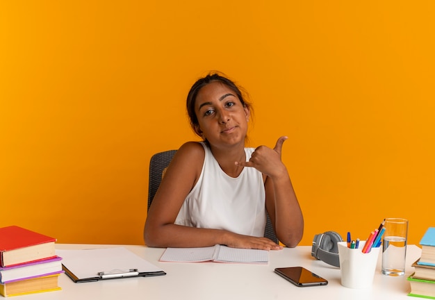 Heureux jeune écolière assis au bureau avec des outils scolaires montrant le geste d'appel téléphonique isolé sur un mur orange