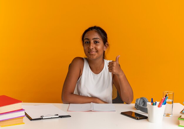 Heureux jeune écolière assis au bureau avec des outils scolaires son pouce vers le haut sur orange