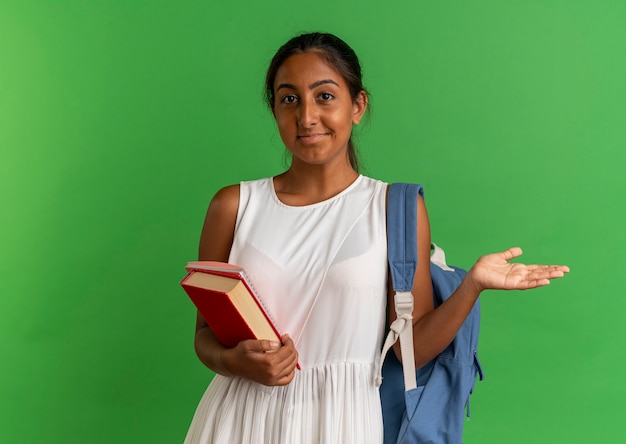 Heureux jeune écolière portant un sac à dos tenant un livre avec un ordinateur portable et répandre la main isolée sur vert