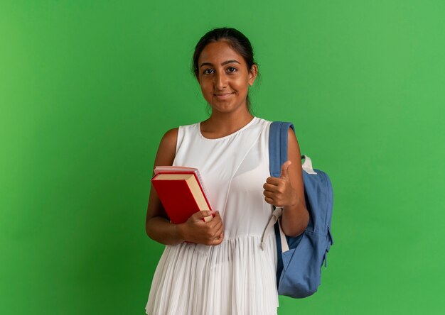 Heureux jeune écolière portant sac à dos tenant livre avec ordinateur portable son pouce vers le haut sur le vert