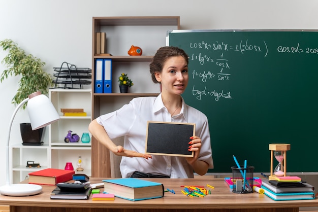 Heureux jeune enseignante de mathématiques assise au bureau avec des fournitures scolaires tenant et pointant sur un mini tableau noir regardant à l'avant en classe