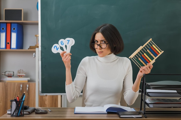 heureux jeune enseignante portant des lunettes tenant un boulier avec un ventilateur numéro assis au bureau avec des outils scolaires en classe