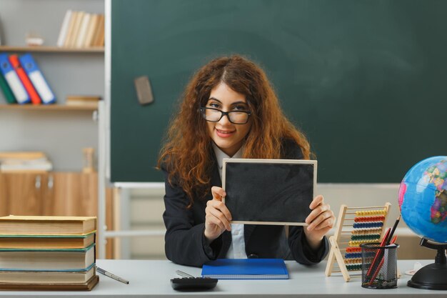 Photo gratuite heureux jeune enseignante portant des lunettes tenant un mini tableau assis au bureau avec des outils scolaires en classe