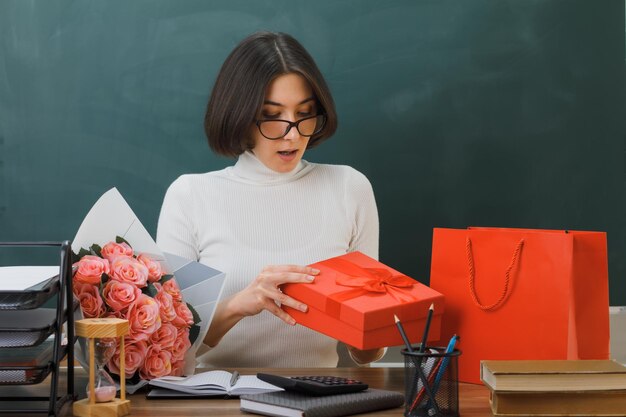 heureux jeune enseignante tenant et regardant présent assis au bureau avec des outils scolaires en classe