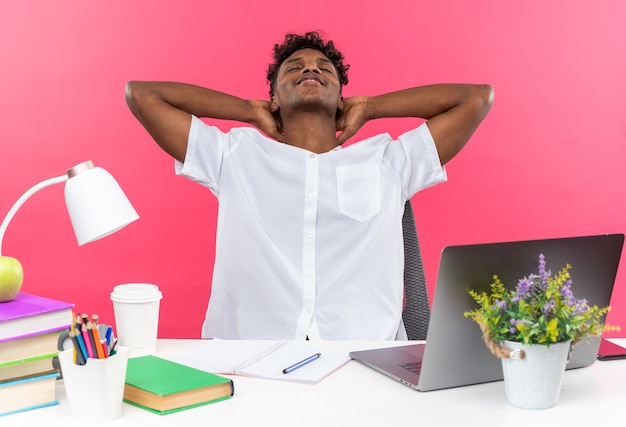 Photo gratuite heureux jeune étudiant afro-américain assis les yeux fermés au bureau avec des outils scolaires mettant les mains sur sa tête isolé sur un mur rose