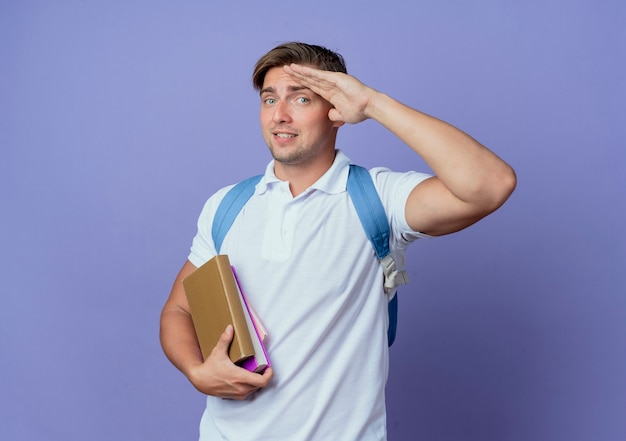 Heureux jeune étudiant beau mâle portant un sac à dos tenant des livres et montrant le geste de salut isolé sur bleu