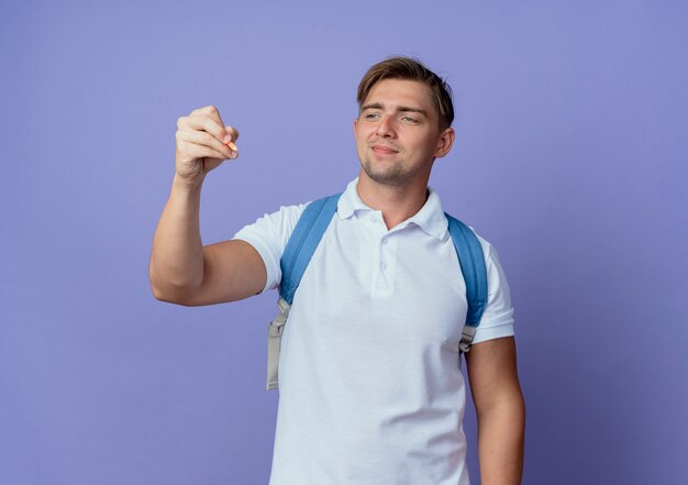 Heureux jeune étudiant beau mâle portant un sac à dos tenant et regardant un stylo isolé sur bleu