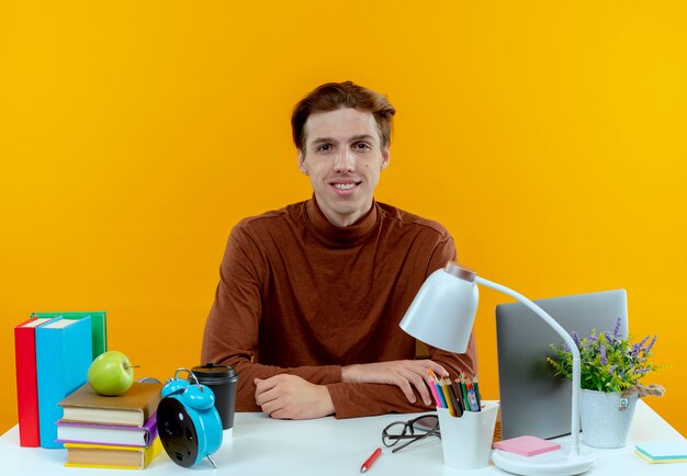 Heureux jeune étudiant garçon assis au bureau avec des outils scolaires sur jaune