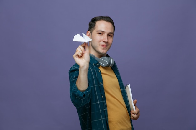 Heureux jeune étudiant de sexe masculin portant des écouteurs autour du cou debout dans la vue de profil tenant un bloc-notes regardant la caméra montrant un avion en papier isolé sur fond violet