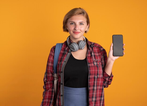 Heureux jeune étudiante slave avec des écouteurs portant un sac à dos tient le téléphone