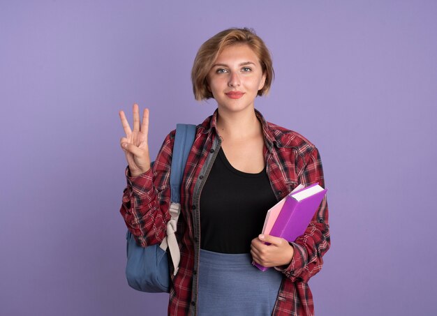 Heureux jeune étudiante slave portant un sac à dos tient des gestes de livre et de cahier trois avec les doigts