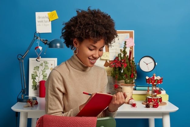 Photo gratuite heureux jeune femme afro-américaine écrit des notes dans le journal, se prépare pour les examens la veille de noël, pose dans la salle d'étude moderne