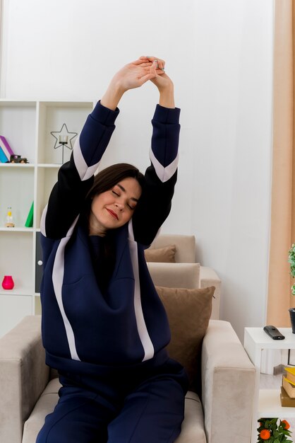 Heureux jeune femme assez caucasienne assise sur un fauteuil dans un salon conçu en gardant les mains ensemble et en s'étirant les yeux fermés