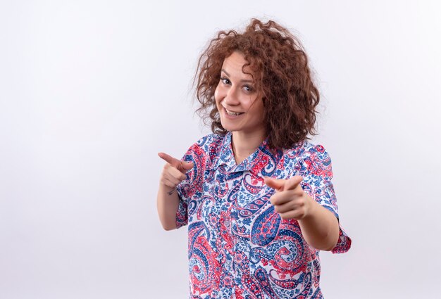 Heureux jeune femme aux cheveux bouclés courts en chemise colorée souriant confiant pointant avec l'index à la caméra