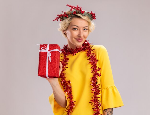 Heureux jeune femme blonde portant une couronne de tête de Noël et guirlande de guirlandes autour du cou tenant un paquet cadeau de Noël regardant la caméra isolée sur fond blanc