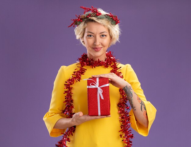 Heureux jeune femme blonde portant une couronne de tête de Noël et guirlande de guirlandes autour du cou tenant un paquet cadeau regardant la caméra isolée sur fond violet