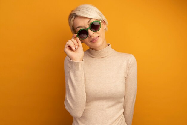 Heureux jeune femme blonde portant et saisissant des lunettes de soleil regardant à l'avant isolé sur un mur orange avec espace de copie