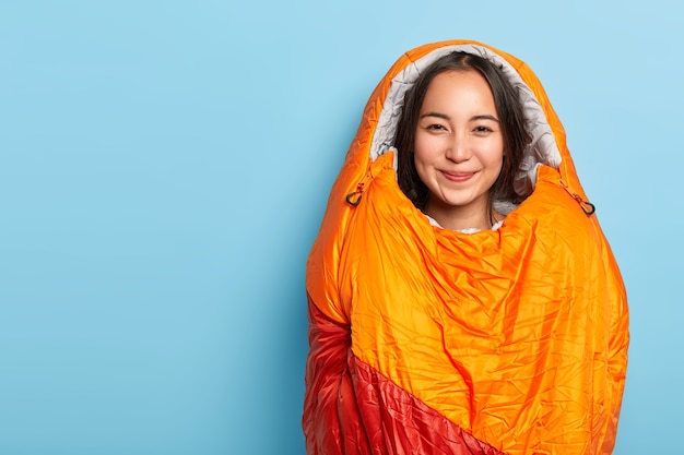 Photo gratuite heureux jeune femme brune asain enveloppé dans un sac de couchage orange chaud, passe du temps libre activement, étant un campeur avtive, se dresse contre le mur bleu