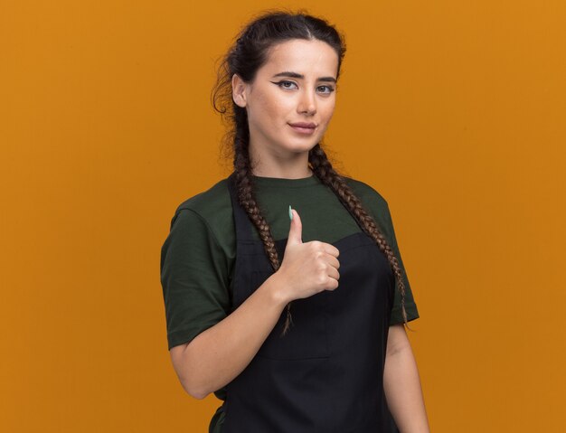 Heureux jeune femme coiffeur en uniforme montrant le pouce vers le haut isolé sur un mur orange