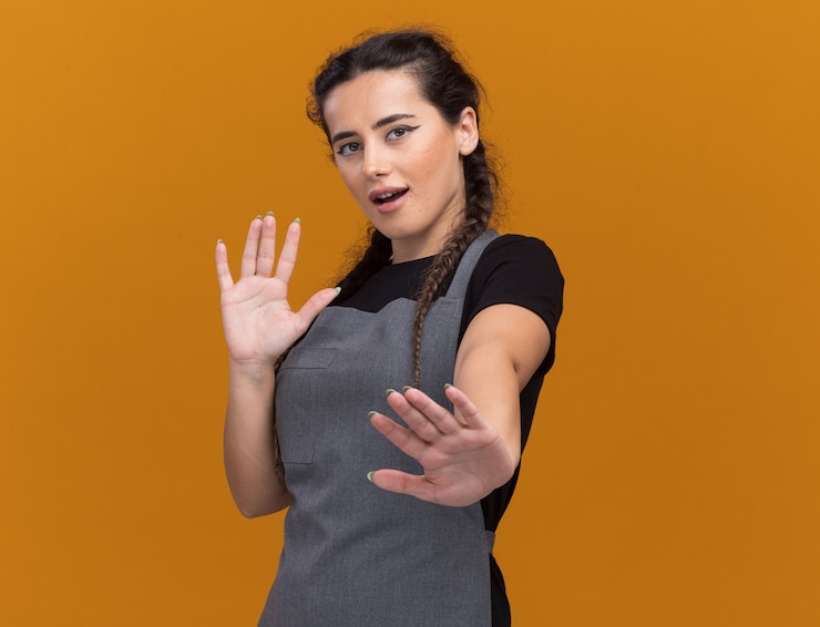 Photo gratuit heureux jeune femme coiffeur en uniforme tenant les mains isolé sur mur orange