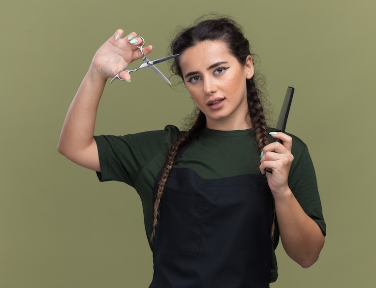 Photo gratuit heureux jeune femme coiffeur en uniforme tenant le peigne et élever des ciseaux isolé sur mur vert olive