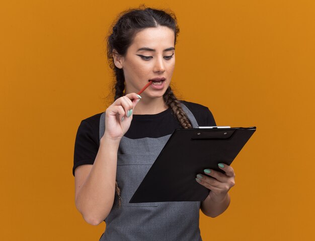 Heureux jeune femme coiffeur en uniforme tenant et regardant le presse-papiers mettre un crayon sur la bouche isolé sur un mur orange