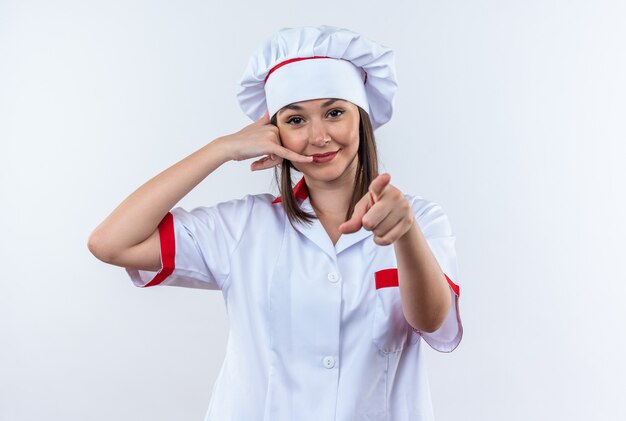 Heureux jeune femme cuisinier portant l'uniforme de chef montrant des points de geste d'appel téléphonique à l'appareil photo isolé sur fond blanc