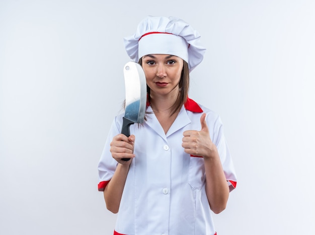 Photo gratuite heureux jeune femme cuisinier portant l'uniforme de chef tenant le couperet montrant le pouce vers le haut isolé sur mur blanc