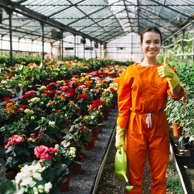 Photo gratuite heureux jeune femme jardinier avec arrosoir gesticulant pouce en l'air