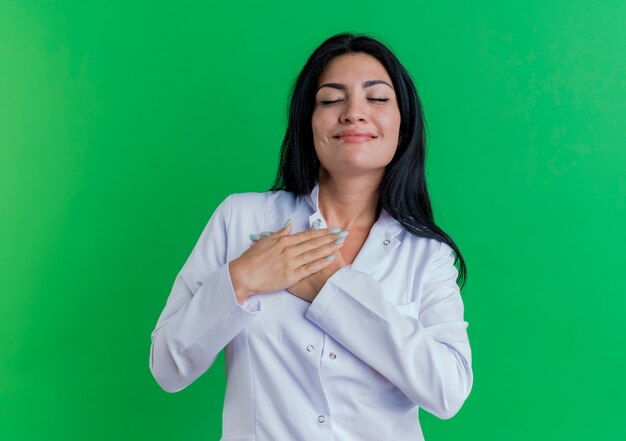 Heureux jeune femme médecin portant une robe médicale mettant les mains sur la poitrine avec les yeux fermés