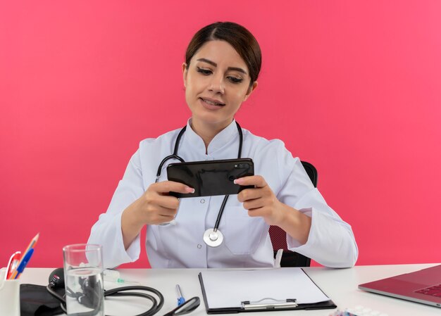 Heureux jeune femme médecin portant une robe médicale et un stéthoscope assis au bureau avec des outils médicaux et un ordinateur portable à l'aide de téléphone mobile isolé sur un mur rose