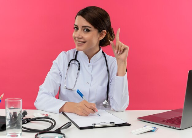 Heureux jeune femme médecin portant une robe médicale avec stéthoscope assis au bureau de travail sur ordinateur avec des outils médicaux écrire quelque chose sur le presse-papiers pointe vers le haut sur le mur rose