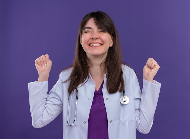 Photo gratuite heureux jeune femme médecin en robe médicale avec stéthoscope garde les poings les yeux fermés sur fond violet isolé avec copie espace