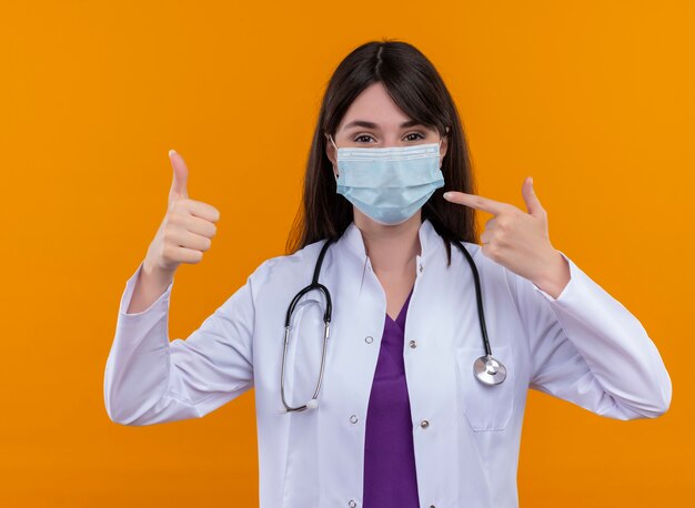 Heureux jeune femme médecin en robe médicale avec stéthoscope porte un masque médical jetable Thumbs up et pointe vers le masque sur fond orange isolé