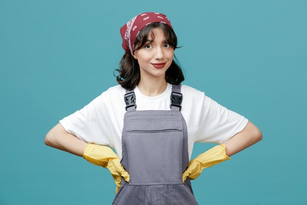 Heureux jeune femme nettoyeur portant un bandana uniforme et des gants en caoutchouc en gardant les mains sur la taille en regardant la caméra isolée sur fond bleu