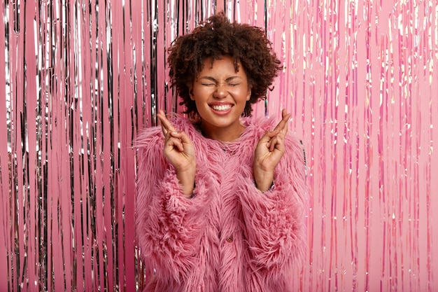 Photo gratuite heureux jeune femme à la peau sombre avec un large sourire, porte un manteau rose à la mode, croise les doigts