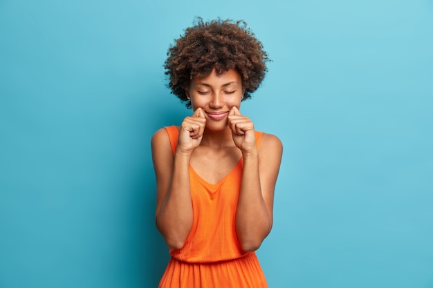 Photo gratuite heureux jeune femme à la peau sombre se tient avec les yeux fermés garde les mains près des joues et des sourires