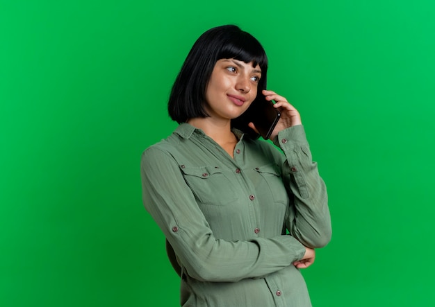 Heureux jeune femme de race blanche brune se tient sur le côté en parlant au téléphone et en regardant côté isolé sur fond vert avec espace de copie