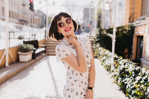 Heureux jeune femme en robe blanche souriant dans la rue. Fille brune aux cheveux courts à lunettes de soleil appréciant le printemps.